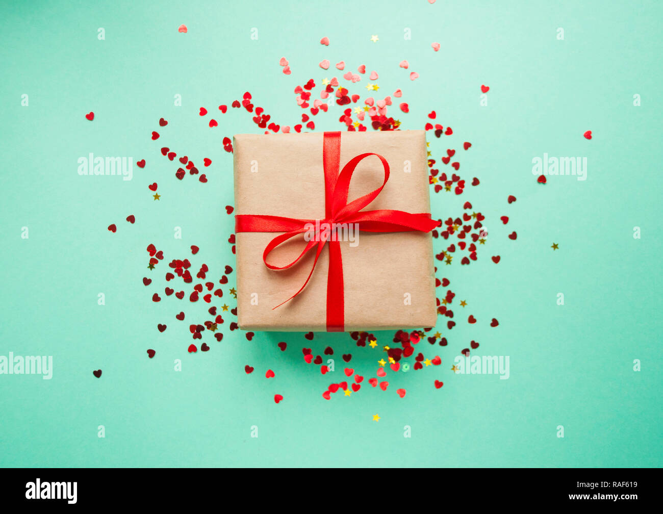 Geschenkbox in braun gefärbte Handwerk Papier gewickelt und mit roter Schleife auf Türkisfarbenem Hintergrund mit rotem Konfetti gebunden. Stockfoto