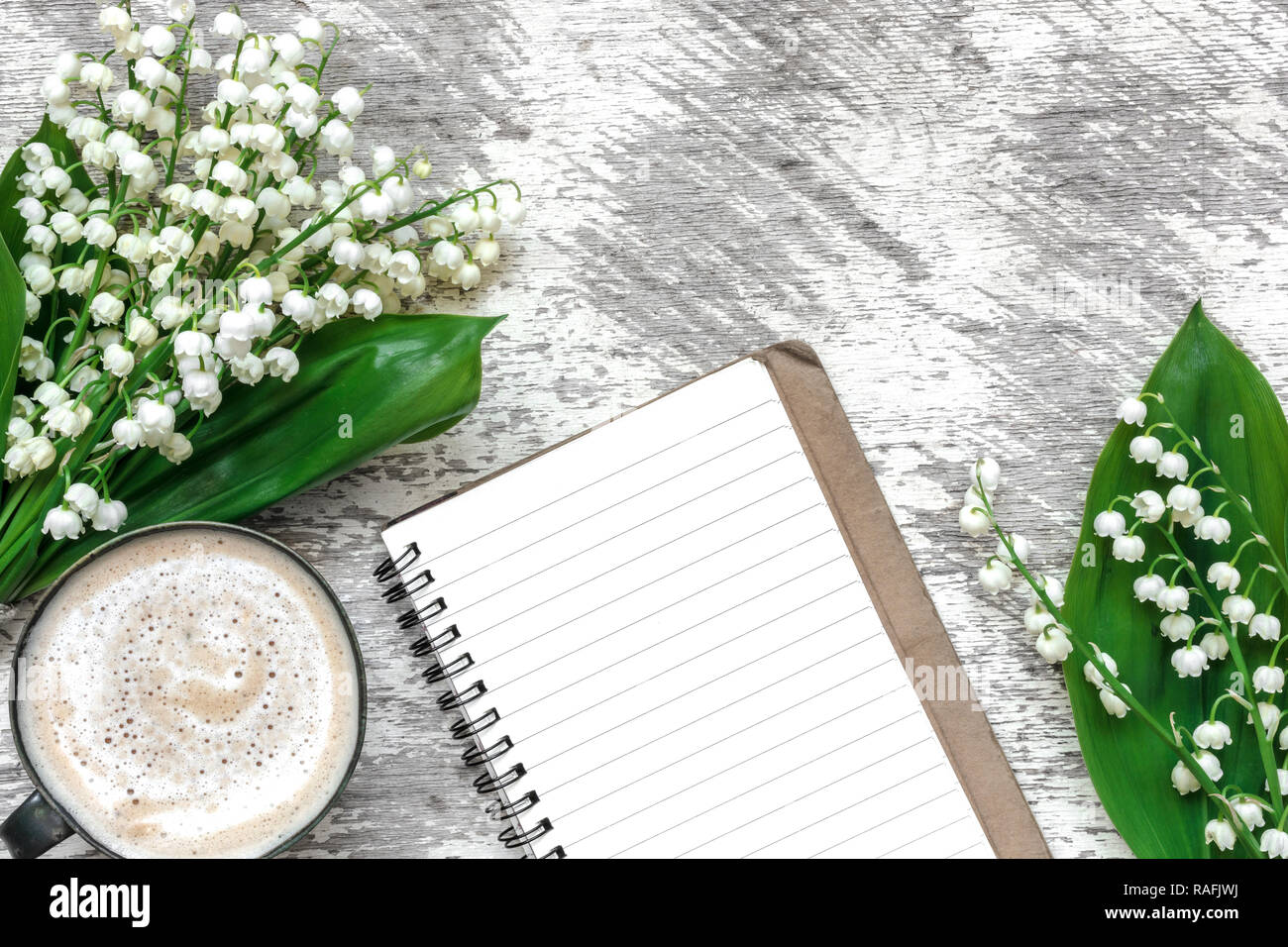 Tasse Cappuccino mit Feder Maiglöckchen Blumen und gesäumt von Notebook über weiße Holztisch. valentines oder Tag der Frauen Hintergrund. nach oben Blick.mo Stockfoto