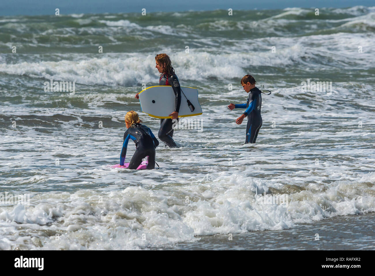 Frau und zwei kleine Jungen stehen am Rande des Wassers über das Meer zu gehen, Bodyboard auf West Wittering Strand, Chichester, West Sussex, UK eingeben Stockfoto