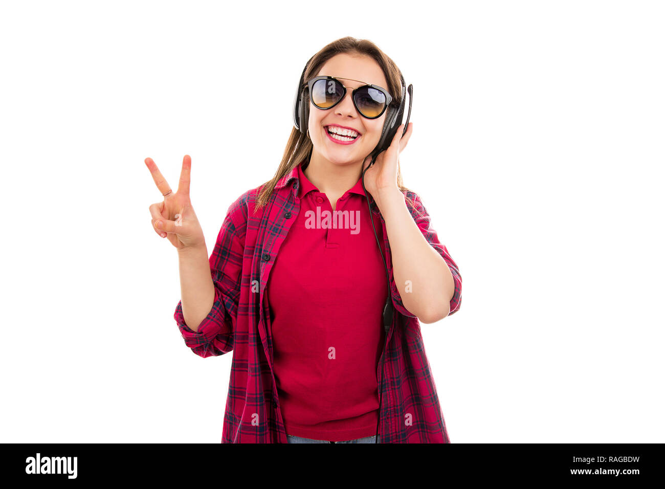 Lächelnd zeitgenössische Teenager Frau in Sonnenbrille Hören von Musik über Kopfhörer und mit zwei Fingern an Kamera Stockfoto