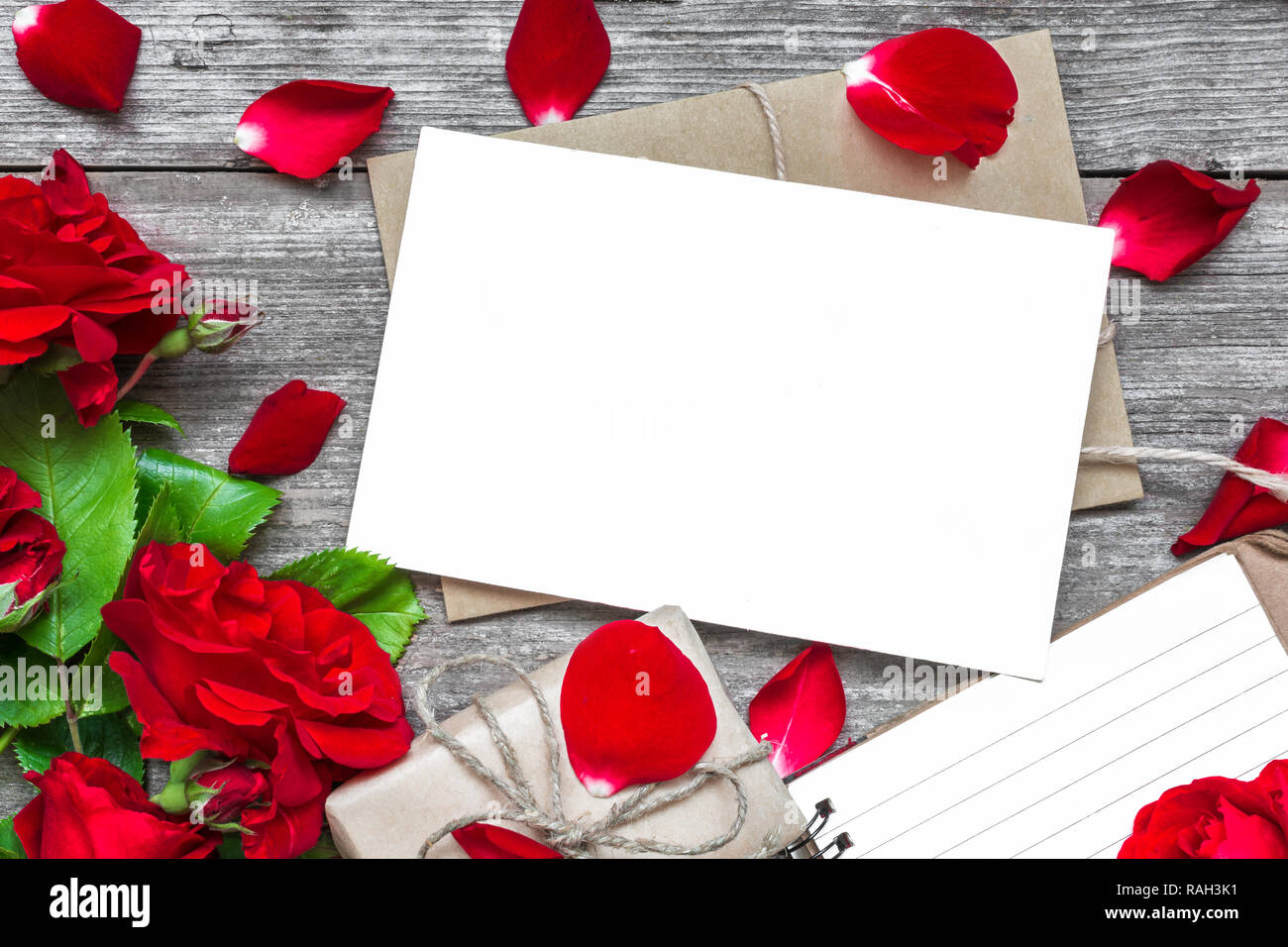 Leere weiße Grußkarte mit rote Rose blumen blumenstrauss und Umschlag mit Blütenblättern, Notebook und Geschenkbox mit rustikalen Holzmöbeln Hintergrund gesäumt. top anzeigen. m Stockfoto
