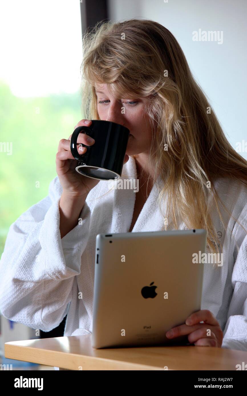 Junge Frau am Morgen in der Küche, im Bademantel und mit einer Tasse Kaffee, Lesen auf einem iPad Tablet-PC Stockfoto