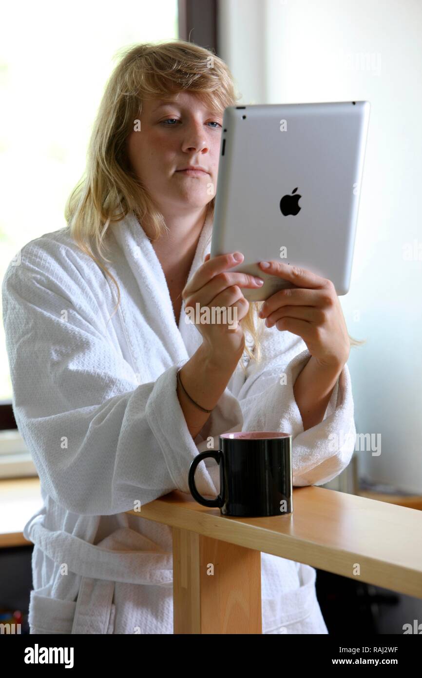 Junge Frau am Morgen in der Küche, im Bademantel und mit einer Tasse Kaffee, Lesen auf einem iPad Tablet-PC Stockfoto
