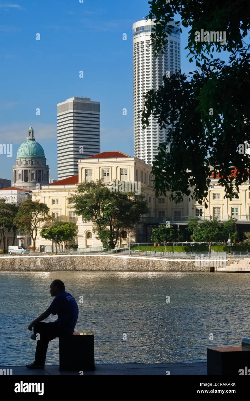 Ein Mann sitzt, der Singapore River in der Nähe von Boat Quay, im Hintergrund die Kaiserin, die Kuppel der National Gallery und Stamford Swissôtel Stockfoto