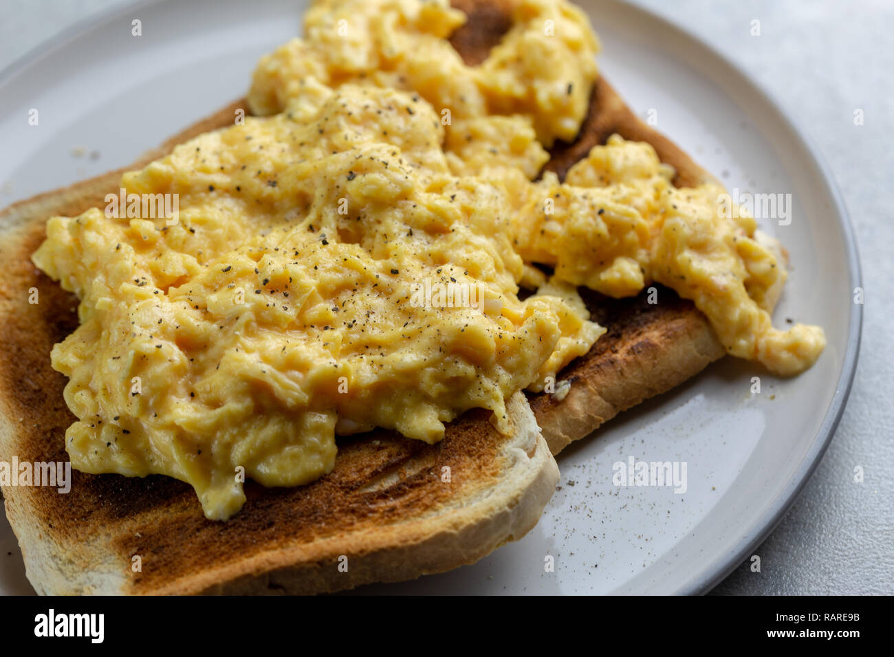 Nahaufnahme von Rührei mit schwarzen Pfeffer auf zwei Scheiben Toast auf eine weiße Platte mit einem grauen Rand auf einer einfachen Grau und Weiß marmorierten Hintergrund Stockfoto