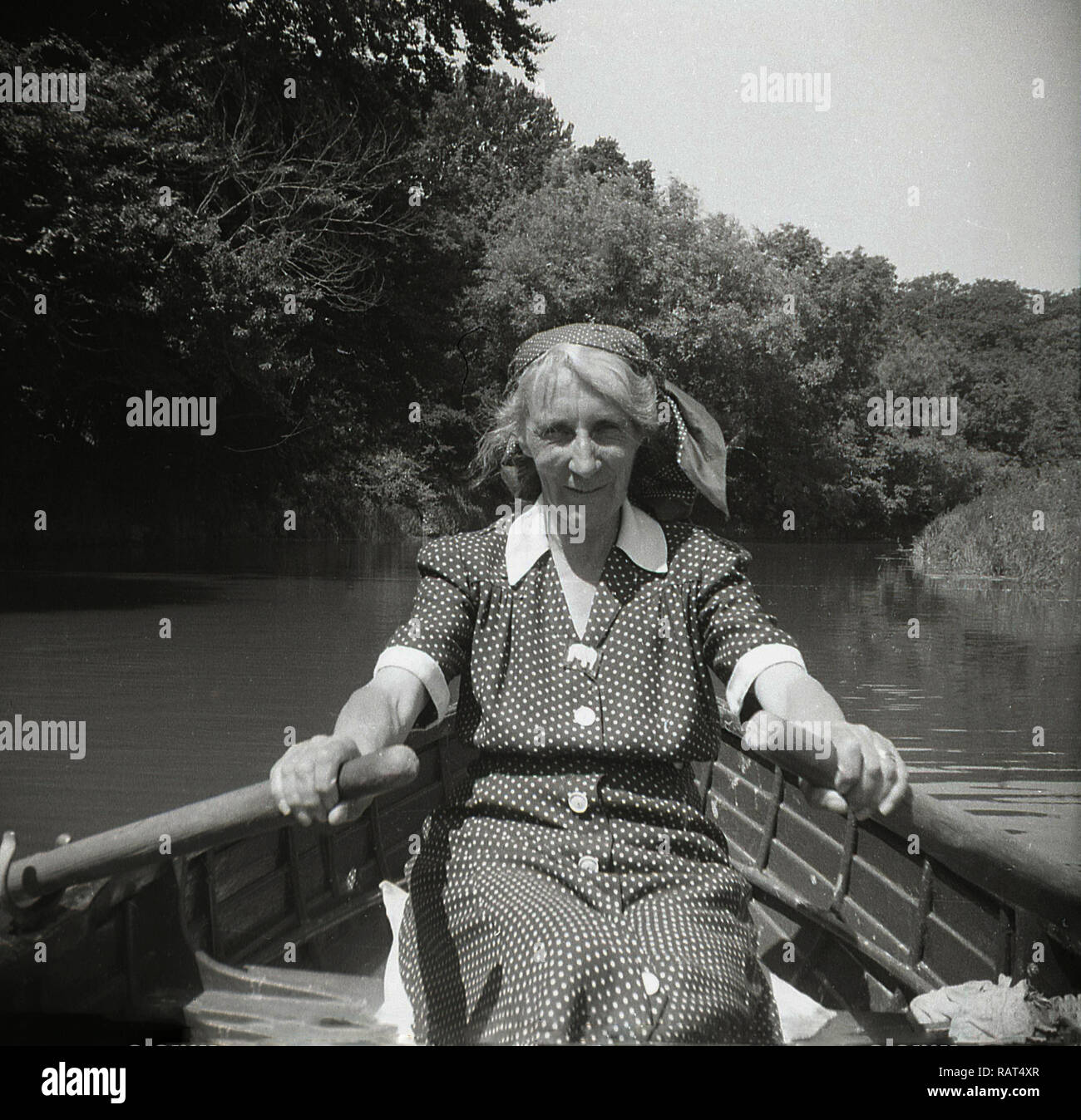 1950, historische, eine Dame mittleren Alters in eine gefleckte Kleid Ruder in der Hand in einem Ruderboot Rudern auf einem Fluss an einem Sommertag, England, Großbritannien sitzen. Stockfoto