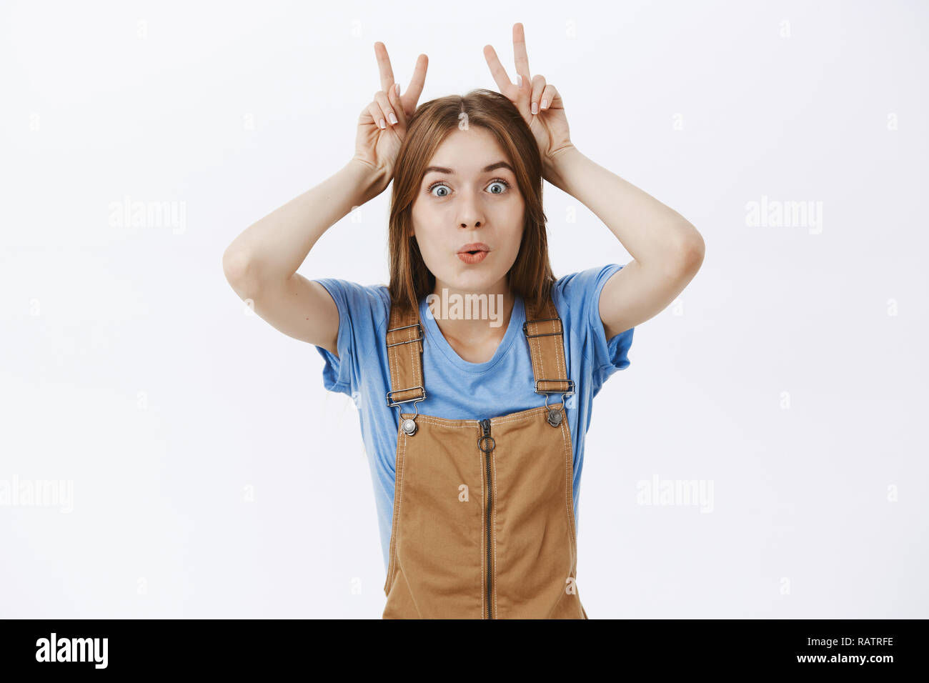 Lustig, verspielt und charismatische junge hübsche Frau in braunen Overalls zeigt Frieden Gesten auf dem Kopf, als Nachahmung Hasenohren falten Lippen und Augen auf der Kamera täuschen um über graue Wand Stockfoto