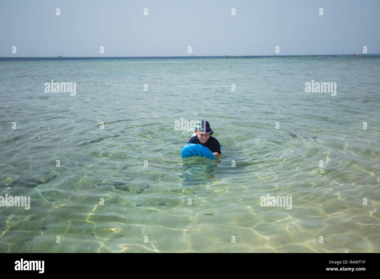 Süße weiß gegerbt Kid im Badeanzug, Kappe und Aqua schuhe Spaß am Sommer, der Sandstrand mit Blau aufblasbares Surfboard. Horizontale Farbfotografie. Stockfoto
