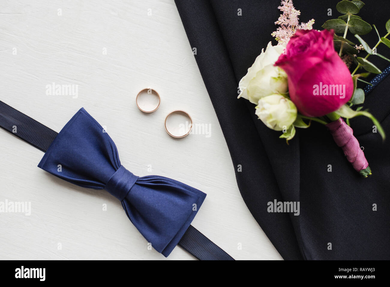 Nahaufnahme, Ansicht von oben in eine elegante, moderne formale männliche Zubehör. Blaue fliege, zwei Goldene Hochzeit Ringe, Blume, Anstecker, schwarze Jacke von Suit festgesteckt. Stockfoto