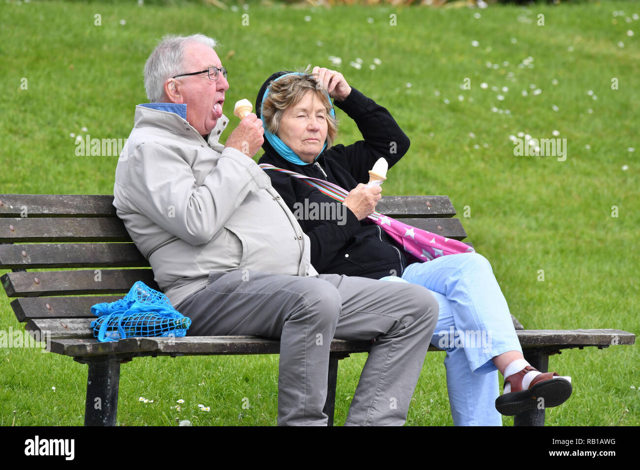 Den mittleren bis älteren Paar Leute sitzen auf einem hölzernen Parkbank tragen Mäntel essen Eis im Alter, an einem kalten Tag im Frühjahr in Großbritannien. Stockfoto