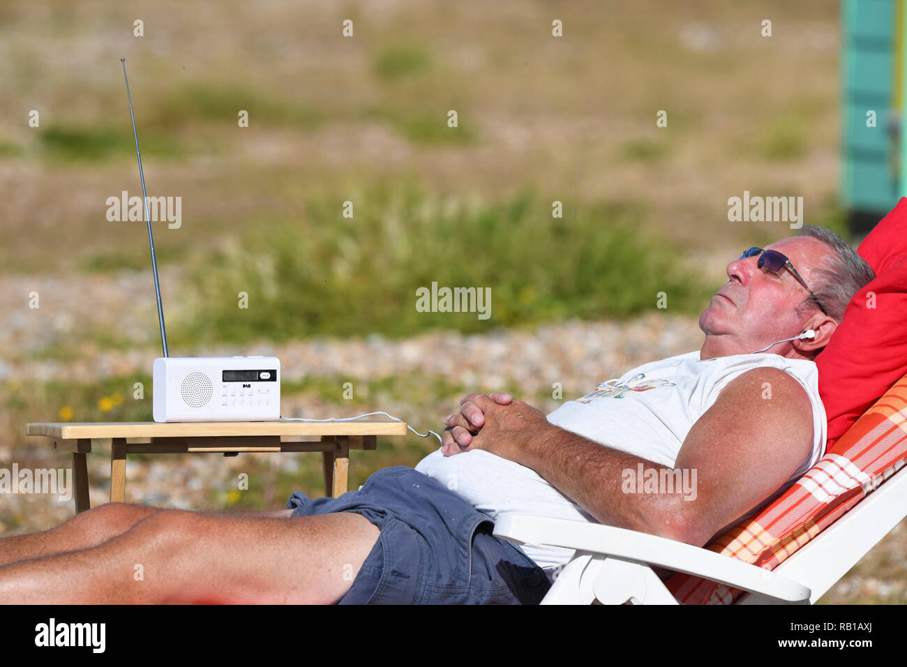 Mann in der Sonne zu liegen in einem Liegestuhl am Strand, beim Tragen der Kopfhörer Radio hören, während der Hitzewelle im Sommer 2018 in Großbritannien. Stockfoto