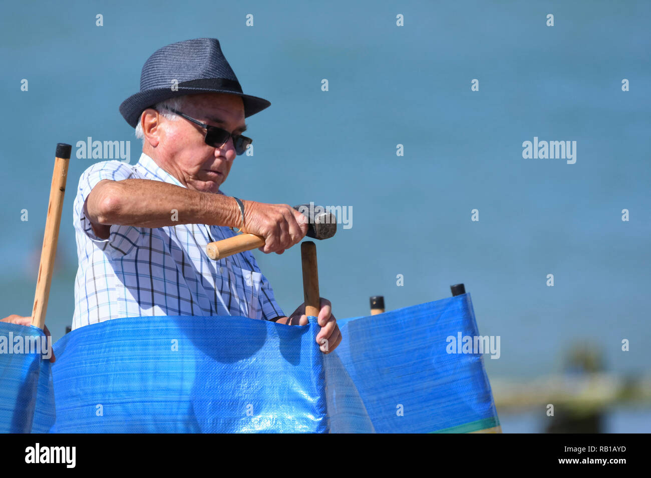 Älterer Mann mit einem Sonnenhut errichten ein Windschutz am Strand im Sommer in Großbritannien. Stockfoto