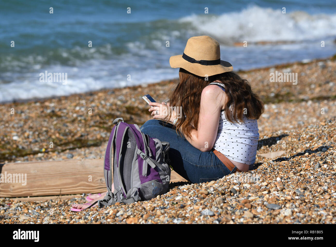 Junge Frau mit Sonnenhut sitzen am Strand mit einem Smartphone an einem heißen Tag im Sommer in Großbritannien. Stockfoto