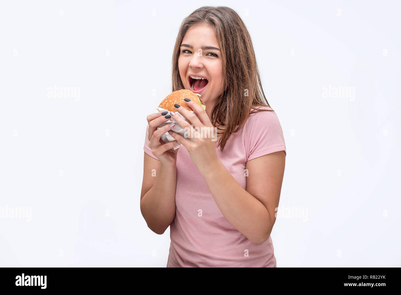 Glückliche junge Frau sehen Sie auf Kamera mit Mund öffnete. Sie halten Burger mit beiden Händen. Auf weissem Hintergrund. Stockfoto