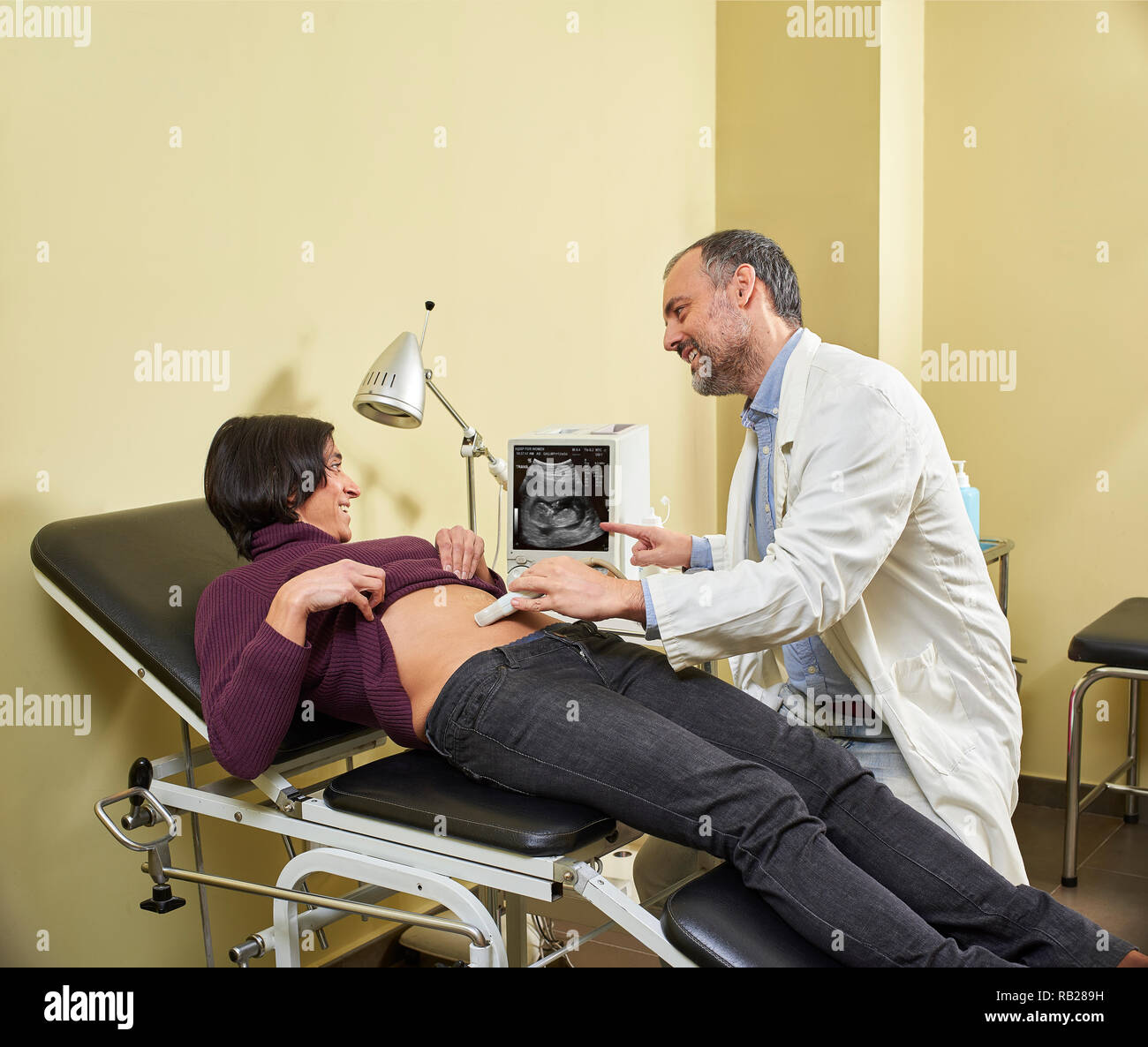 Ein lächelndes Gynäkologen ein Ultraschall Verfahren auf eine glückliche fortyish Hispanic schwangere Frau mit einer medizinischen Ultraschall Scanner scannen. Stockfoto