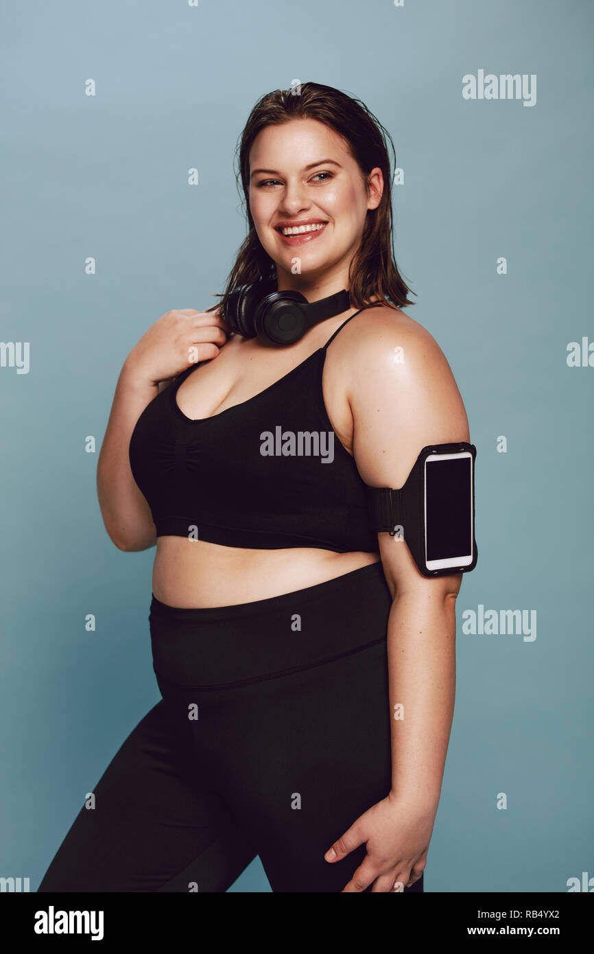 Plus Size junge Dame in Sportbekleidung. Portrait von Fitness junge Frau auf grauem Hintergrund posiert mit Handy am Armband und Kopfhörer um Ihr n Stockfoto