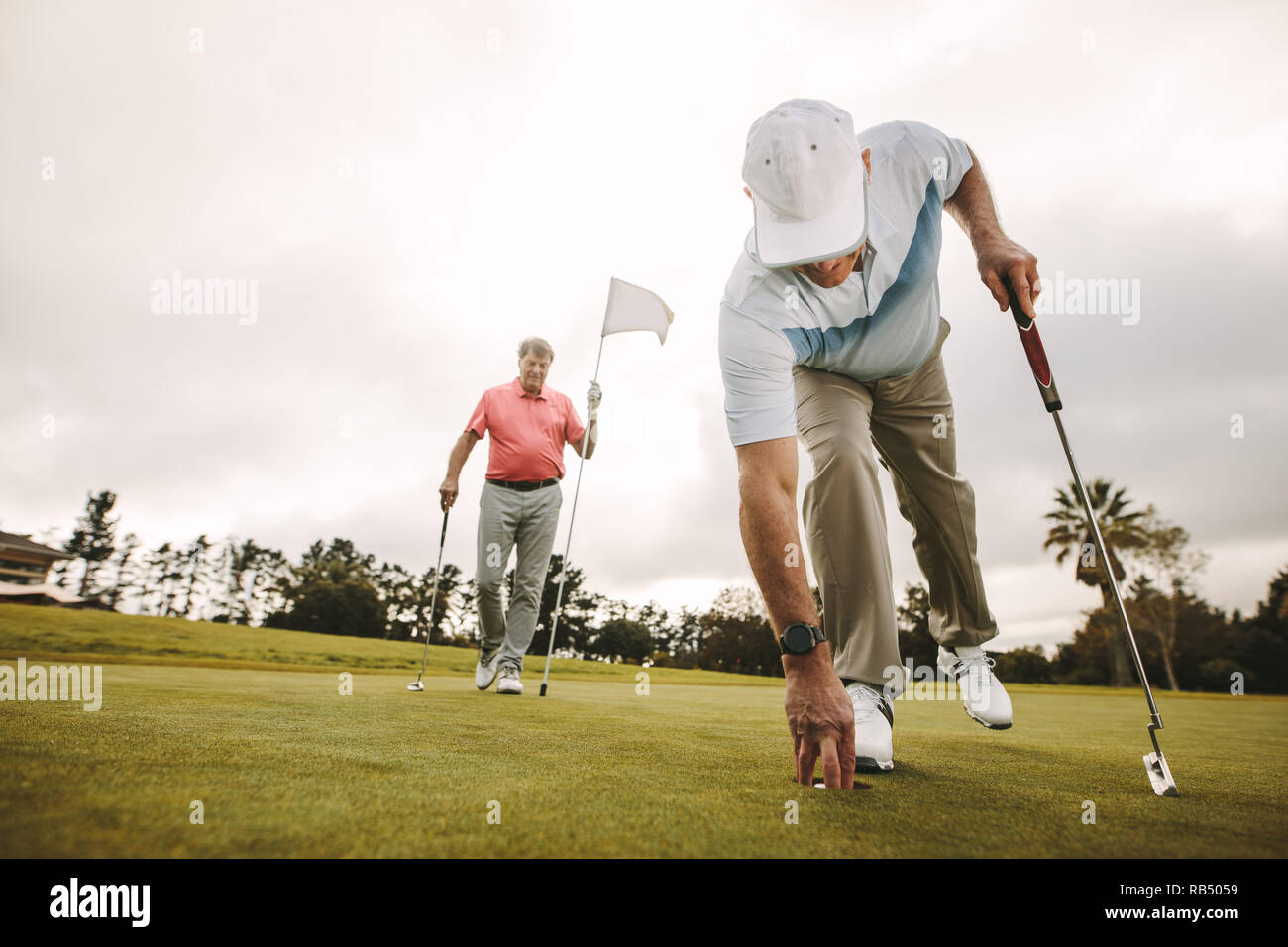 Ältere Golf player Kommissionierung Golf Ball aus der Bohrung mit einem zweiten Spieler im Hintergrund halten die Flagge. Ältere Golf Spieler ein Spiel spielen auf dem Golfplatz Stockfoto
