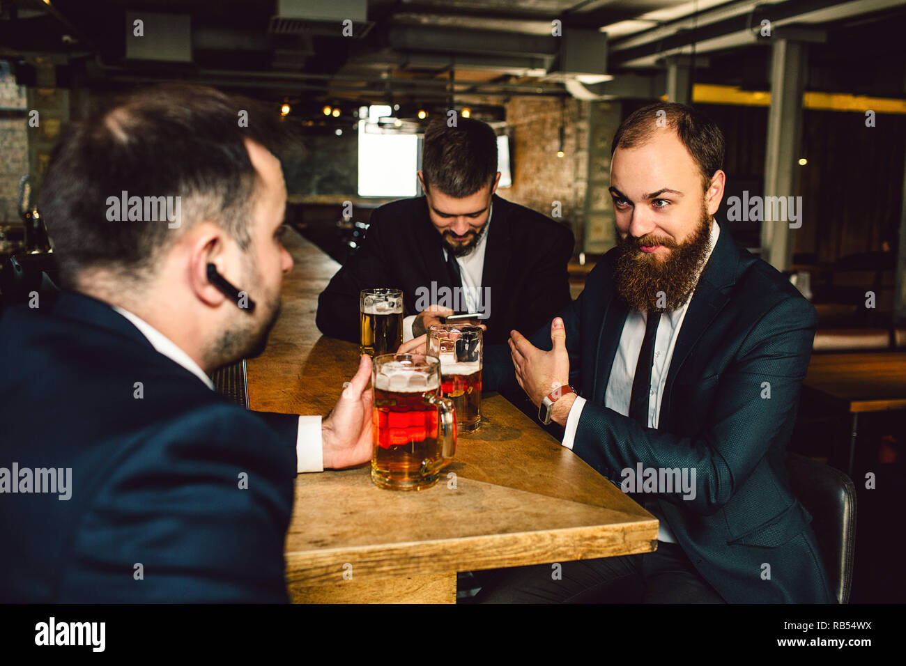 Positive junge bsinessmen in Anzügen sitzen zusammen am Tisch. Sie halten Becher Bier. Kerl vor hat Schwarz In-Ear-Kopfhörer. Männer sind in der Bar. Stockfoto