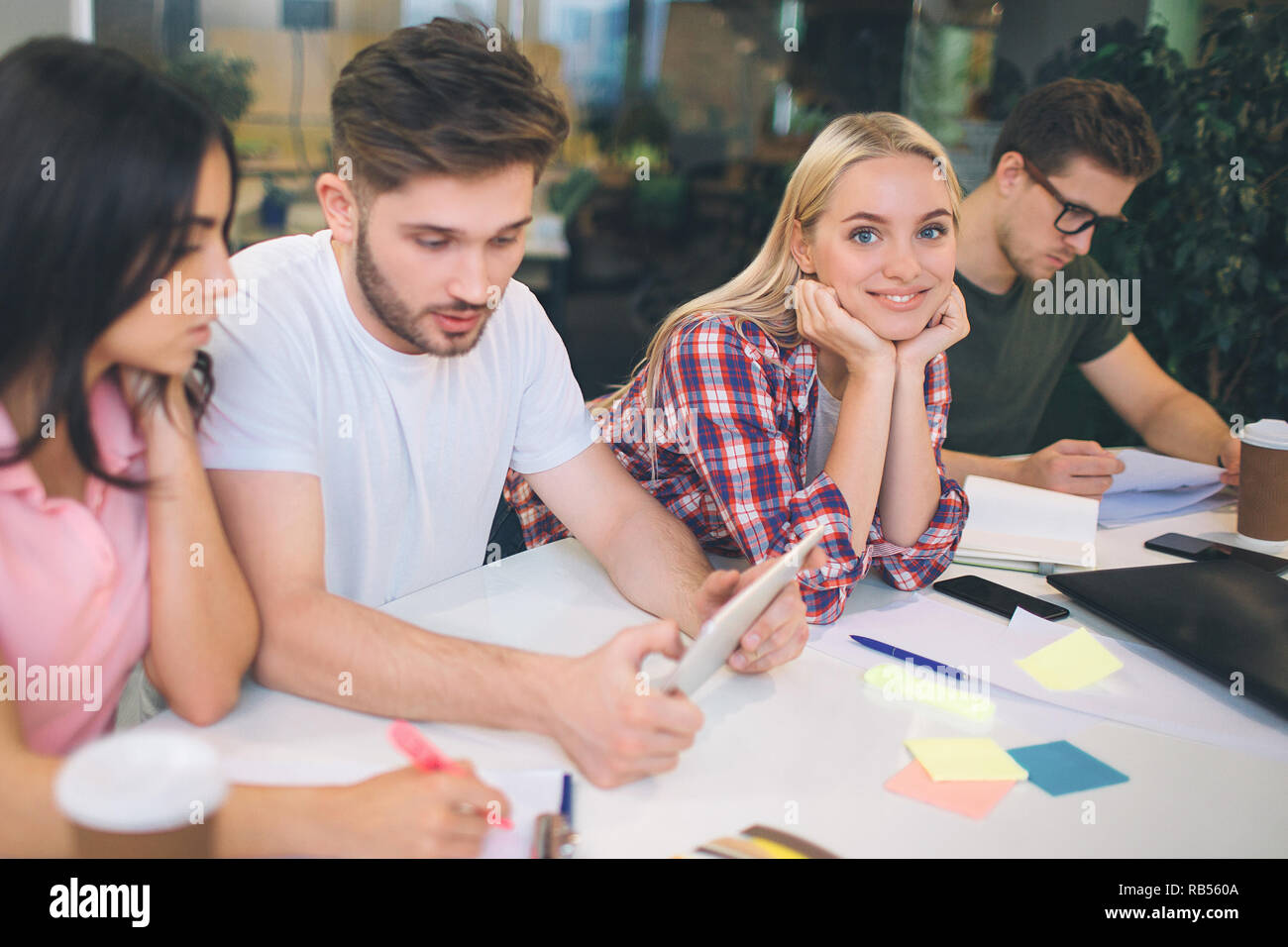 Drei junge peoople Arbeit ernst am Tisch. Sie schauen nach unten. Junge blonde Frau Blick auf Kamera und Lächeln. Sie sitzt zwischen den anderen Studenten. Alle sind sie im selben Zimmer. Stockfoto
