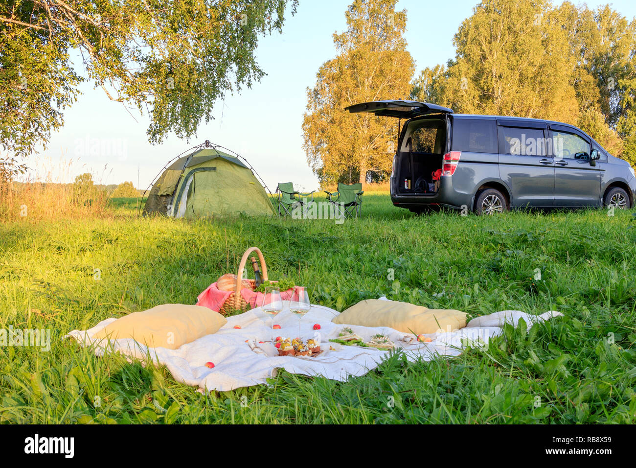 Picknick im Wald, Zelt und Camp Stühle, grau Minibus mit offener Tür, Decke, Weidenkorb, Weingläser, Snacks, Obst, Brot, Kissen. Konzept Stockfoto