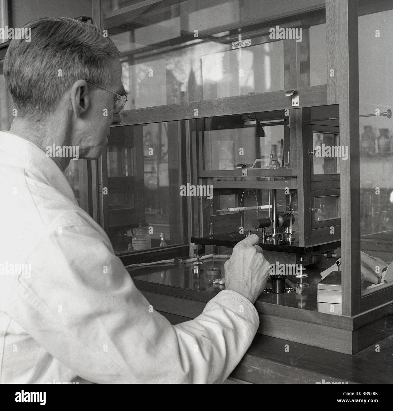 1950, historische, ein reifer Mann Professor in einem weißen Mantel in einer Science Laboratory an der Universität von Oxford, England, ein laborexperiment mit Instrumenten in einem Holz gerahmt Protective Glass Cabinet. Stockfoto