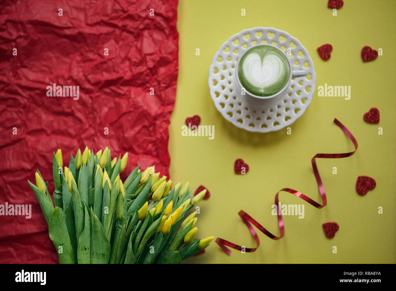 Eine Tasse mit einem leckeren und aromatischen morgen Matcha Latte Kaffee auf einer festlichen Hintergrund mit vielen Herzen. Weiter ist ein Strauß gelber Tulpen. Stockfoto