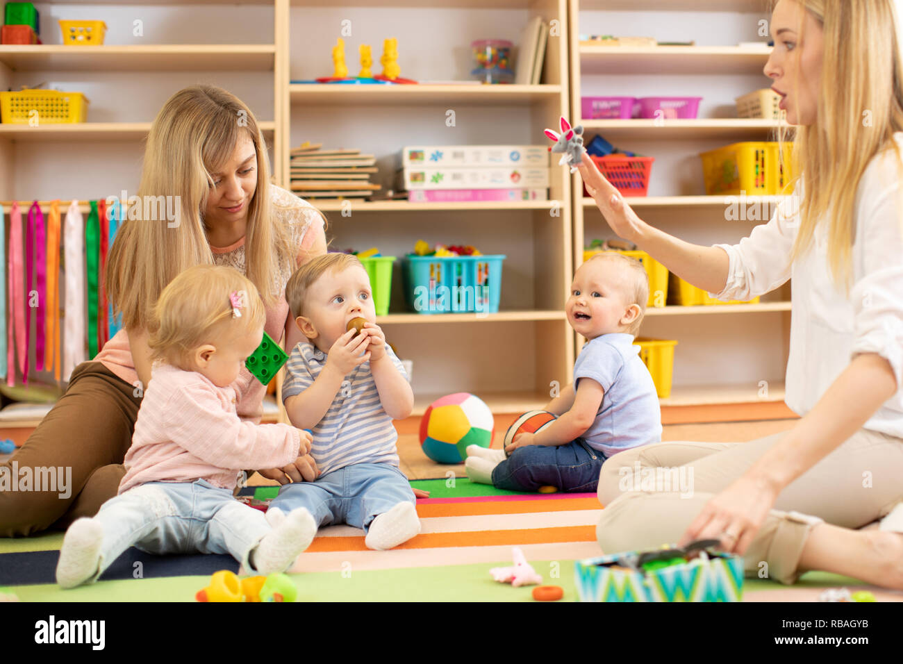 Gruppe von Babys im Kinderzimmer. Kindergärtnerin und Assistent spielen mit Kindern auf gemütlichen Teppich Stockfoto