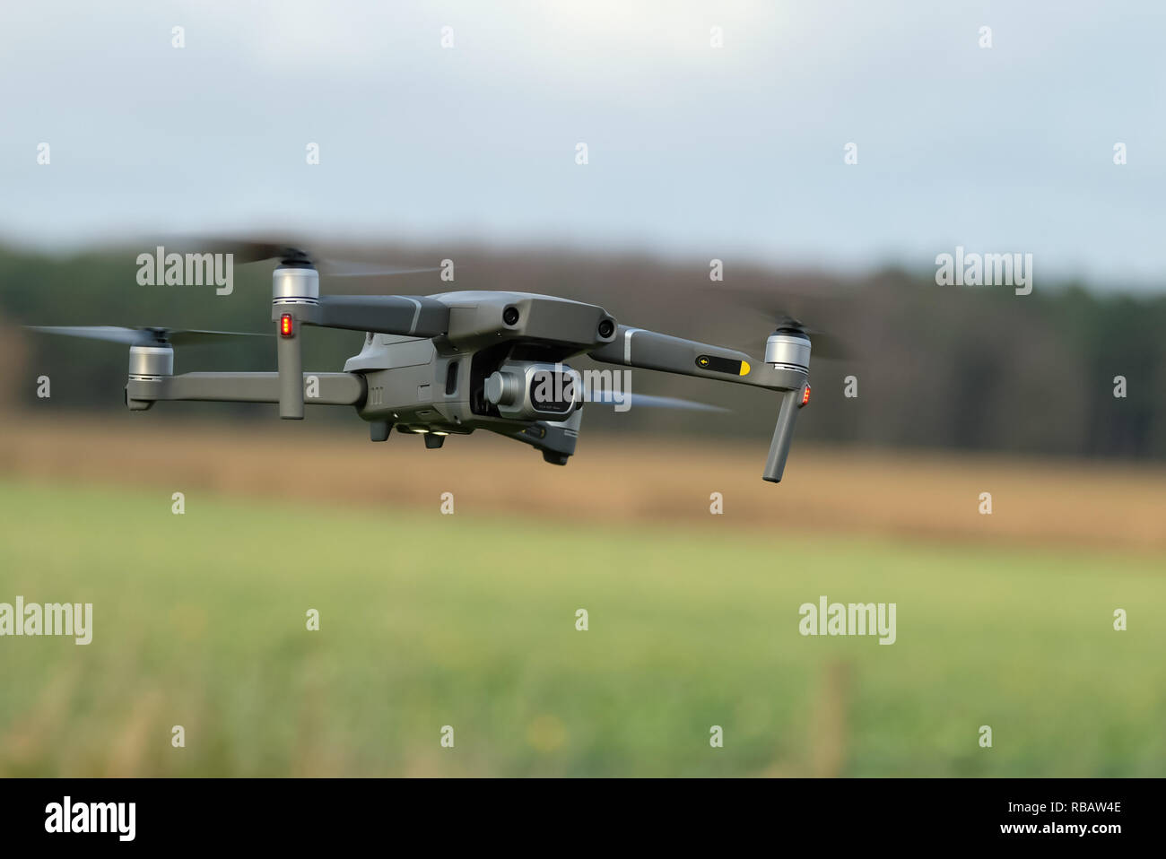 Eine DJI Mavic 2 Pro Quadcopter-Drohne mit montierter Kamera fliegt tief über einem grasbewachsenen Feld, mit einer unscharfen Baumlinie und einem bewölkten Himmel im Hintergrund Stockfoto