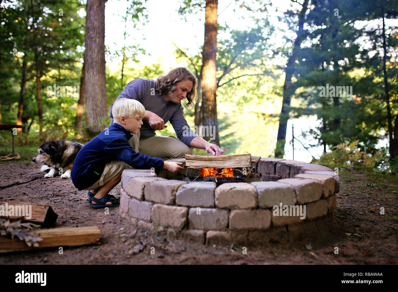 Ein Vater und sein kleiner Sohn beginnt ein Lagerfeuer in einem Feuerring auf einem Campingplatz mit Blick auf einen See im Wald, als ihre älteren Hund in der Nähe legt. Stockfoto