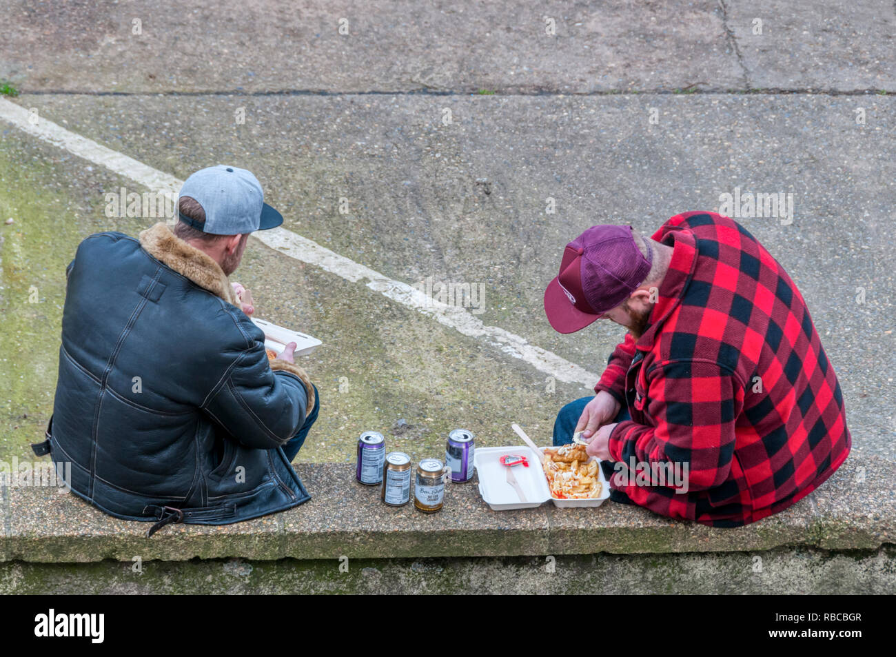 Zwei Männer sitzen auf einer niedrigen Mauer außerhalb essen Fisch und Chips und trinken Dosen von Bitter shandy in Wells-next-the-Sea, Norfolk. Stockfoto