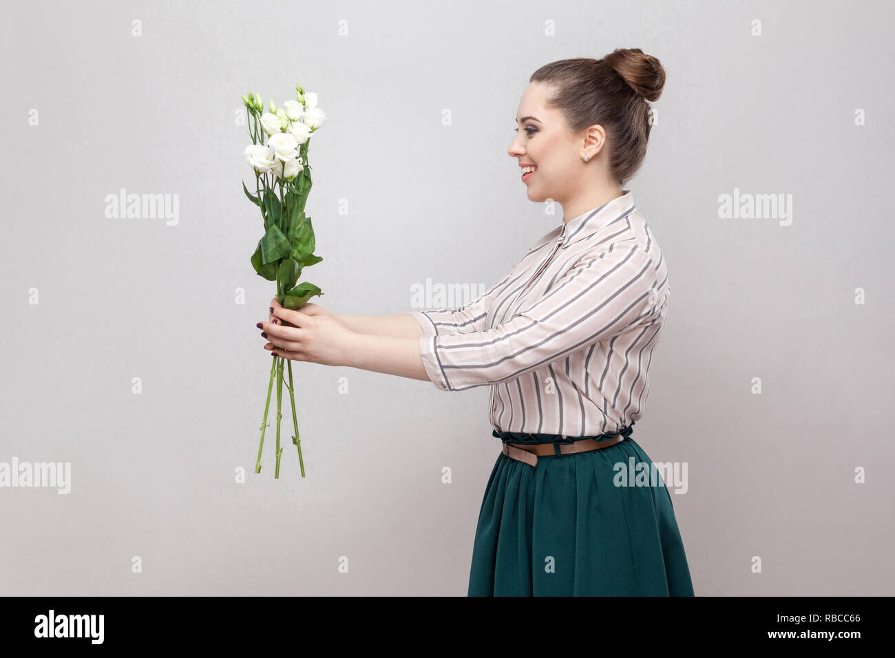 Seite Profil ansehen attraktiver romantische junge Frau im gestreiften Hemd und grün Rock holding Blumenstrauß aus den weißen Blüten und berät Sie. Innen-, Studio Stockfoto
