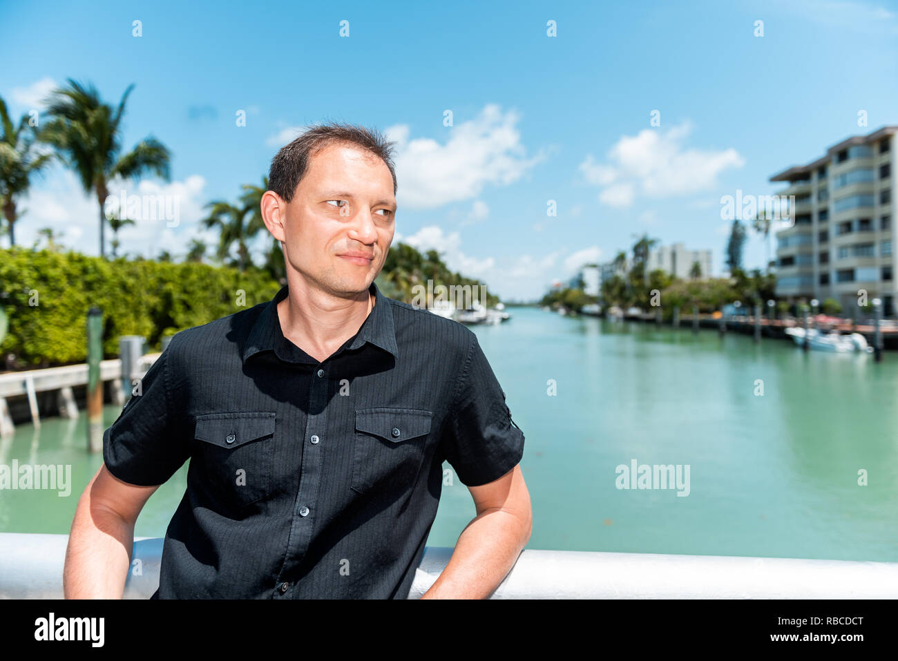 Junge Mann Gesicht mit schwarzen T-Shirt steht auf Brücke Geländer in Bal Harbour, Miami Florida lehnte sich mit Green Ocean Biscayne Bay. Stockfoto