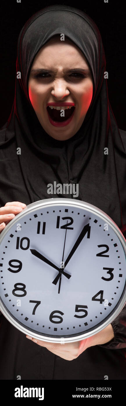 Porträt der schönen schweren Angst junge muslimische Frau mit schwarzen Hijab halten Sie clock in ihre Hände als Laufzeit Konzept auf schwarzem Hintergrund Angst Stockfoto