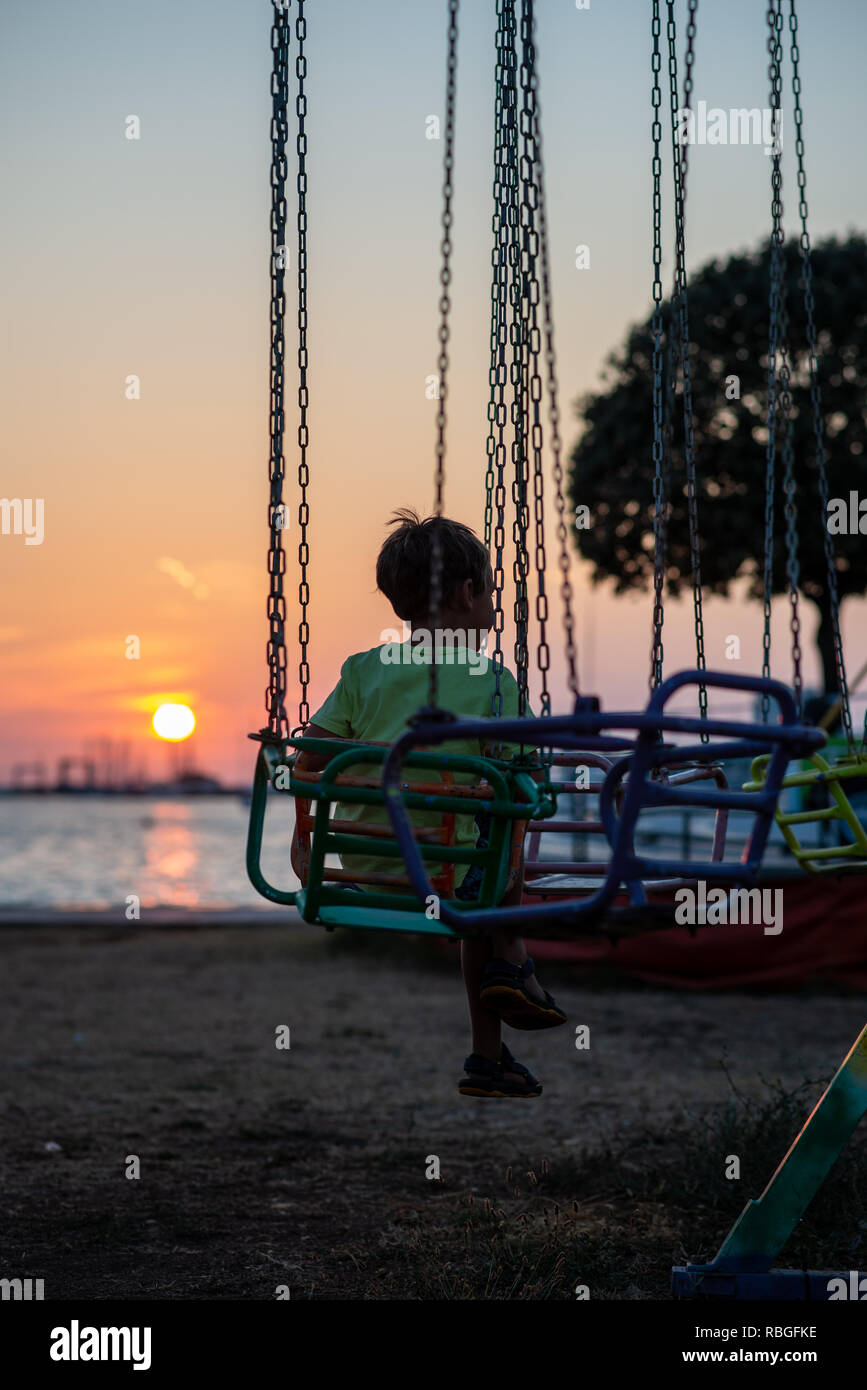 Kleinkind Junge in ein Karussell mit wunderschönen Sonnenuntergang und das Meer im Hintergrund sitzen. Stockfoto