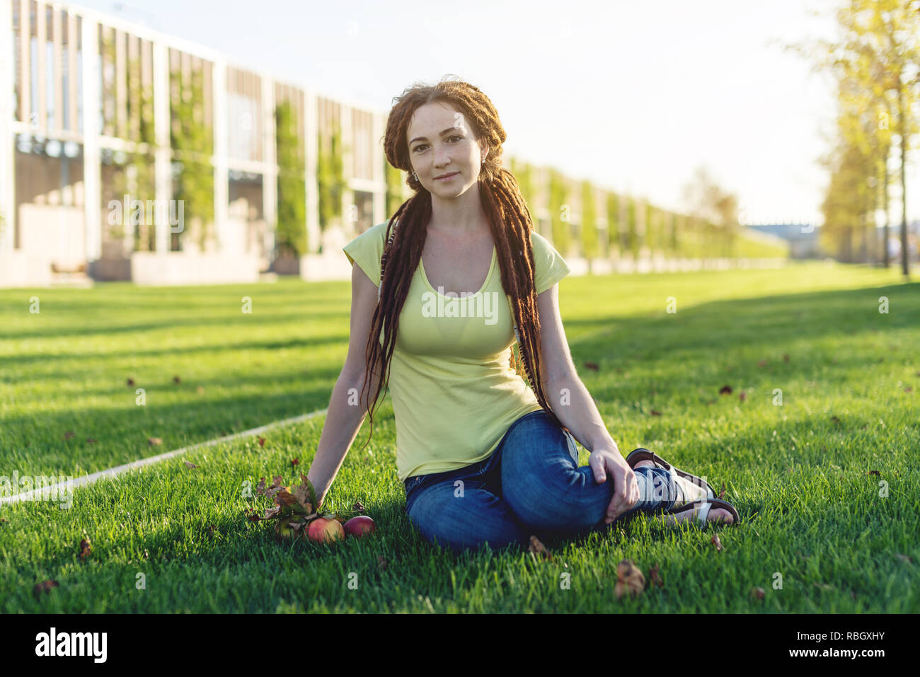 Moderne junge Mädchen mit einer Frisur dreadlocks in den Park auf der grünen Wiese essen Äpfel und geniessen Sie den Herbst sonnige Stimmung Stockfoto