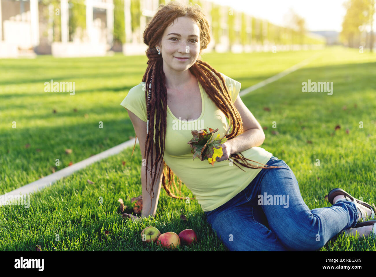 Moderne junge Mädchen mit einer Frisur dreadlocks in den Park auf der grünen Wiese essen Äpfel und geniessen Sie den Herbst sonnige Stimmung Stockfoto