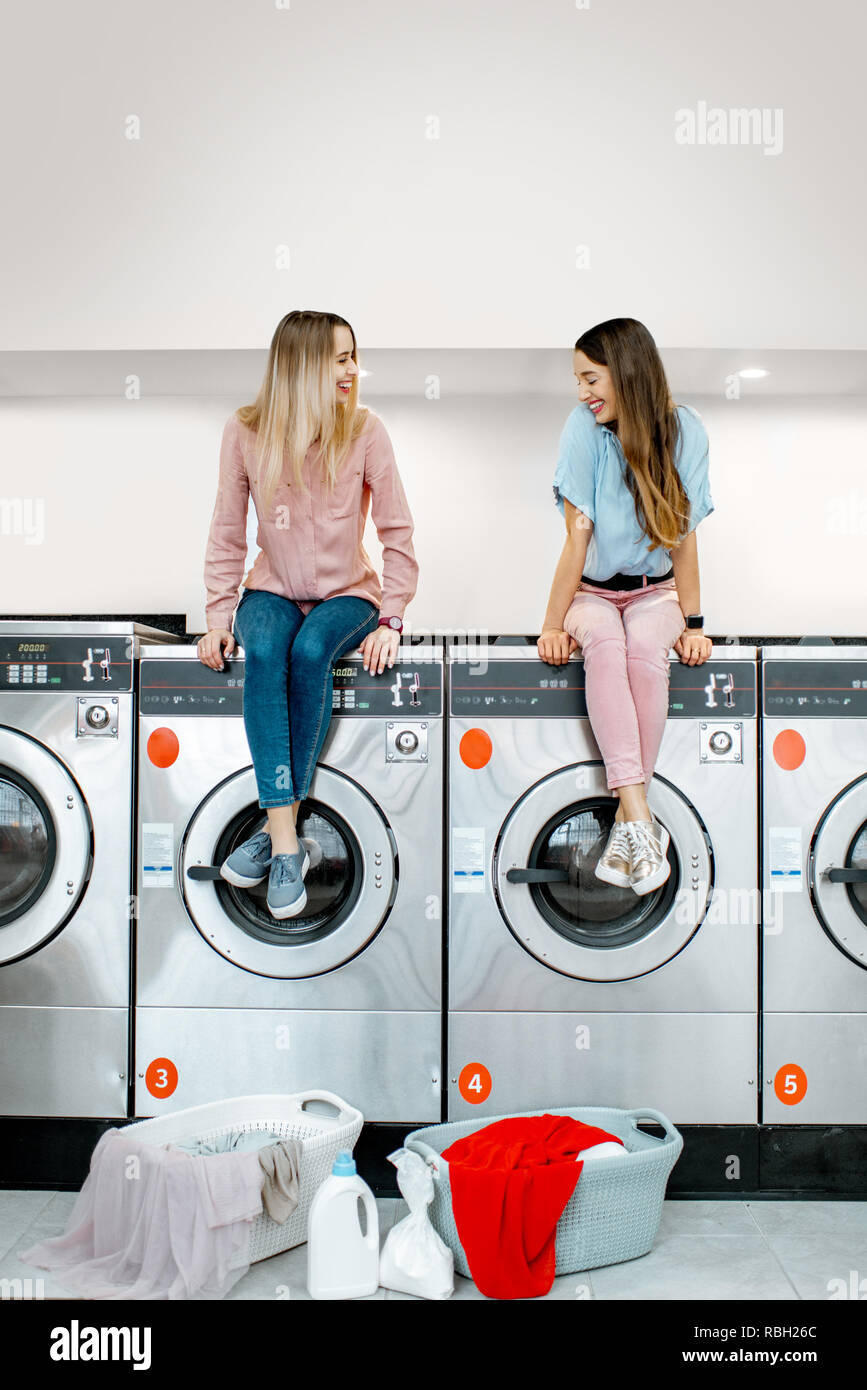 Zwei fröhliche Freundinnen zum Waschen während der Sitzung auf die Waschmaschinen in den Waschsalon mit Selbstbedienung warten Stockfoto