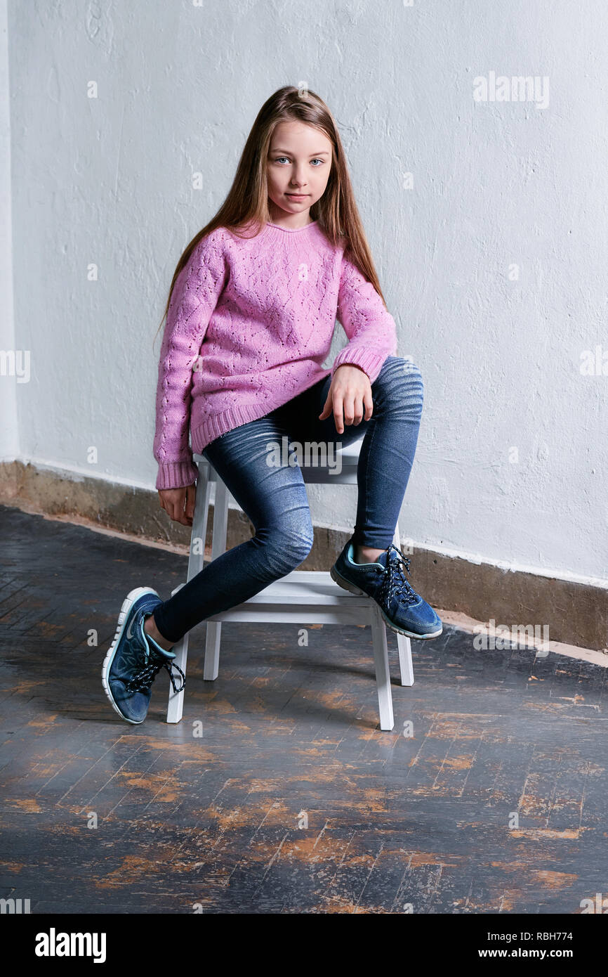 Zuversichtlich Kind mädchen mode Pose sitzt auf einem Stuhl, konkrete weißen Hintergrund. Elegantes Modell in rosa Pullover. Schöne glamourösen Kind Jugendlicher in Kleidung casual, urbanen Stil. Studio gedreht. Stockfoto