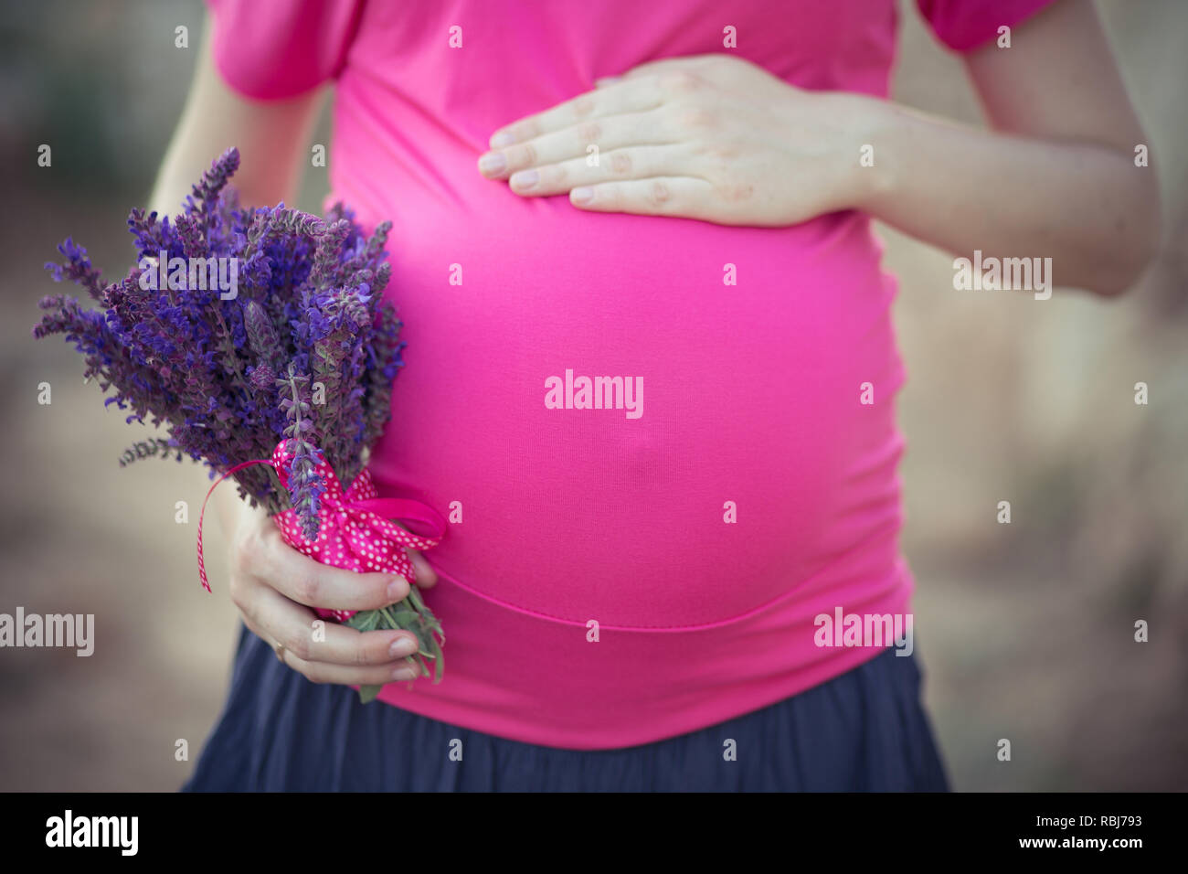 Hübsche junge schwangere Frau Frau mit riesigen wunderschönen grünen Augen und Schmollmund rote Lippen tragen dunkel rosa kleid mit blauen rock, Hände auf ihren Bauch Stockfoto