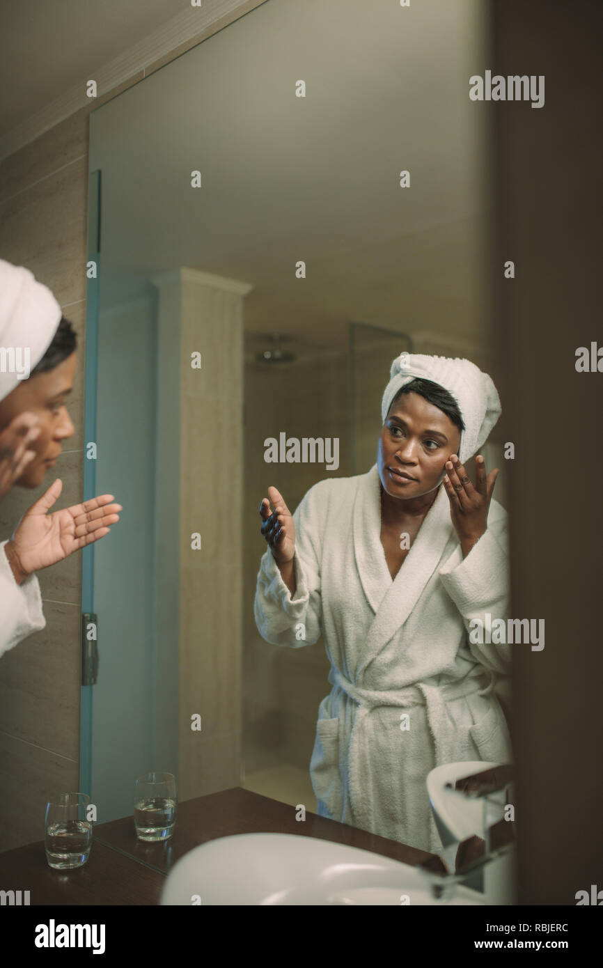 Afrikanische Frau im Bademantel Auftragen kosmetischer Lotion Pflege der Haut im Bad zu nehmen. Face Care Concept. Reflexion der reifen Frau in der mirr Stockfoto
