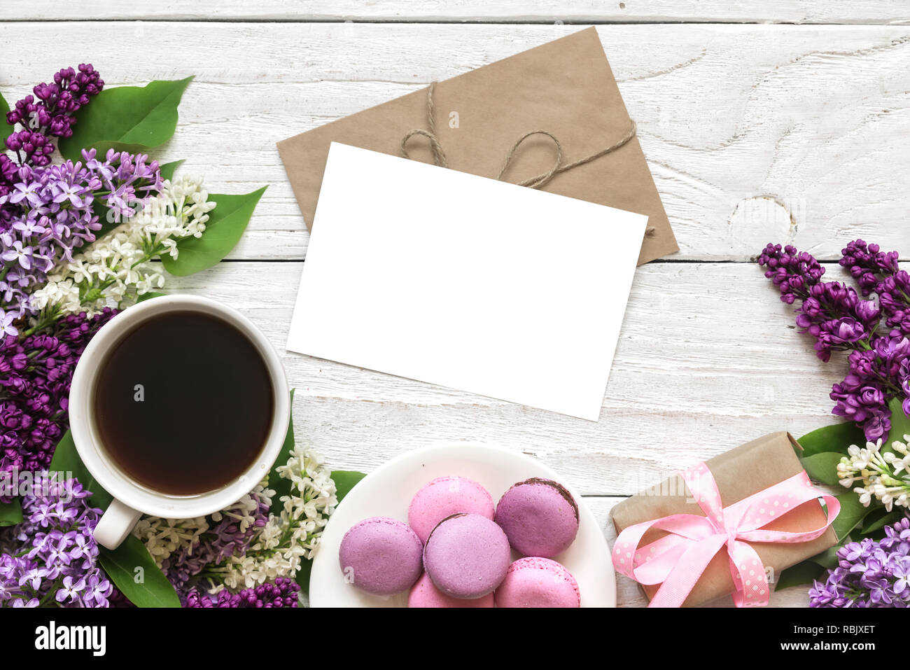 Leere weiße Grußkarte mit lila Blumen, Kaffeetasse, süße Macarons und Geschenkbox auf weißem Holz- Hintergrund. Ansicht von oben mit der Kopie Raum. Mock up Stockfoto