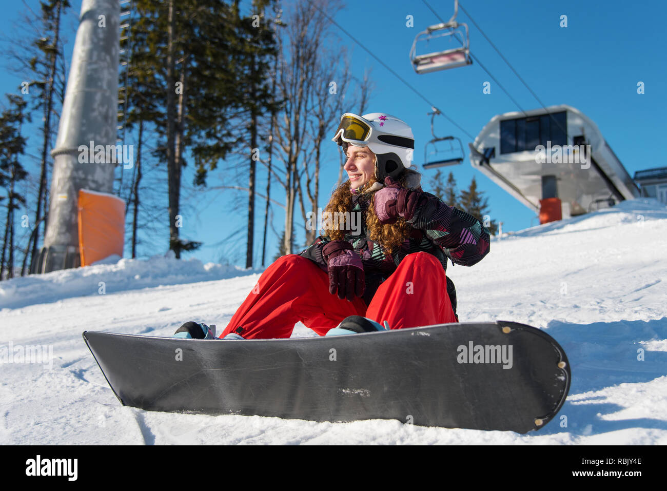 Lächelnde Mädchen mit Snowboard sitzen auf dem Schnee unter einem Skilift  im Skigebiet und Daumen hoch Gesten des guten Klasse Stockfotografie - Alamy