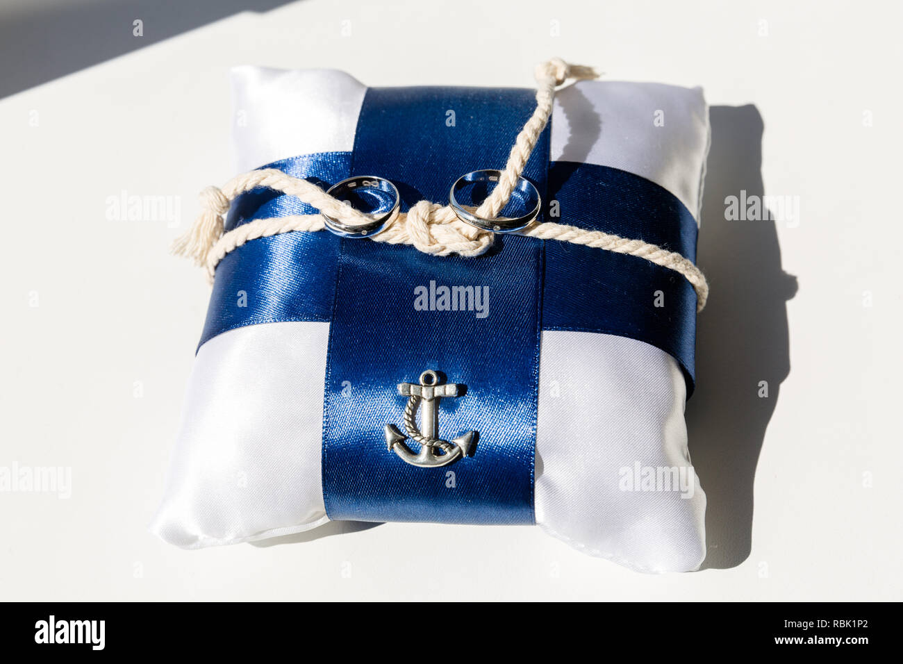 Ein paar Trauringe auf einem weißen Kissen mit blauen Bändern. In der Mitte des Kissens ist ein Seil mit Knoten gebunden. Auf der Seite der Kissen ist ein Metall Dekoration in der Form eines Sea Anchor Stockfoto