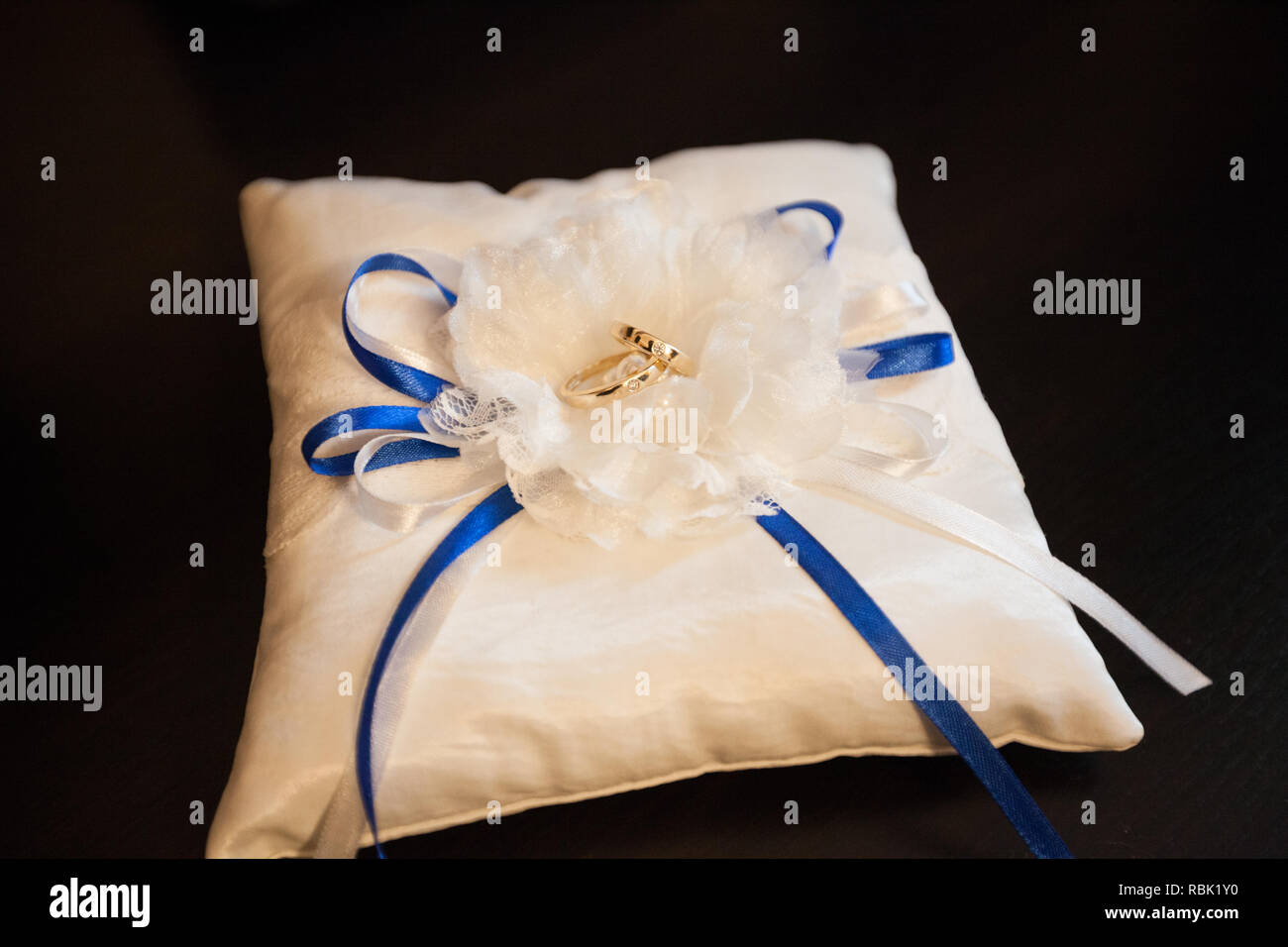 Ein paar Trauringe auf einem weißen Kissen. In der Mitte des Kissens ist ein Bogen aus weißen und blauen Bändern. Kissen auf einem schwarzen Hintergrund. Stockfoto