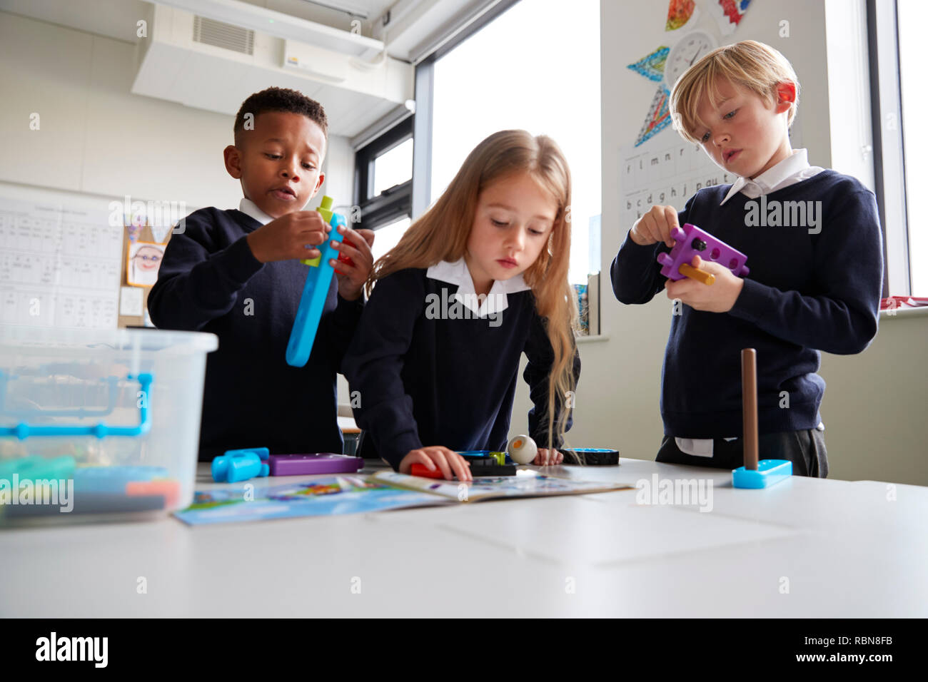 Drei Grundschüler gemeinsam mit Spielzeug Bau Blöcke in einem Klassenzimmer, das Mädchen, das Lesen der Anleitung aus einem Buch, Nahaufnahme, niedrigen Winkel Stockfoto