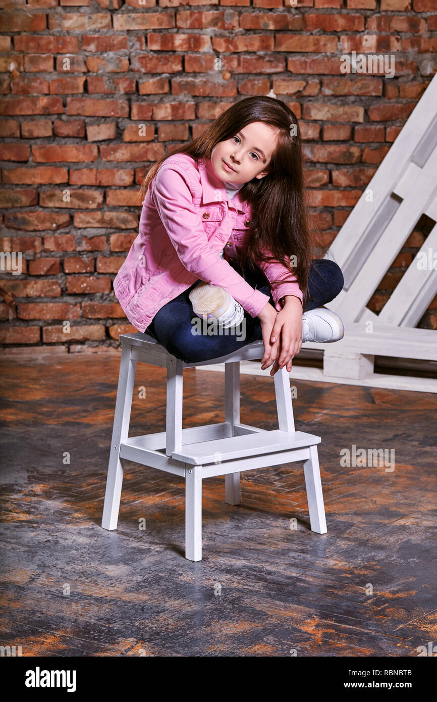 Stilvolle Modell Kind. Portrait Mädchen junge Teenager pose sitzen auf Stuhl. Schöne Brünette wirbt für legere Kleidung, Jugend style Trend. Studio shot, loft Interieur. Stockfoto