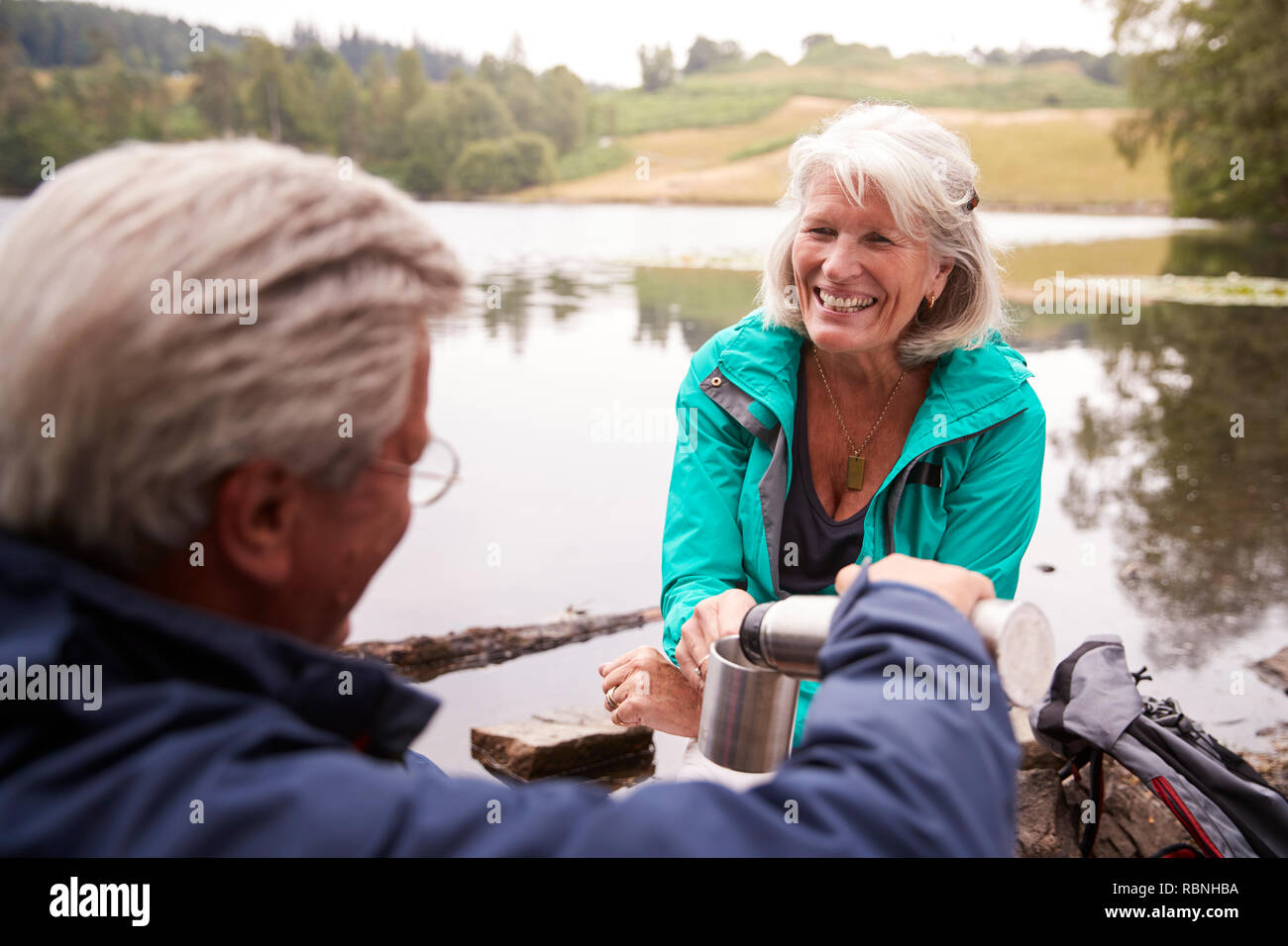 Senior Paar an einem See, mann Kaffee in die Tasse seiner Frau gießen, über die Schulter sehen, Lake District, Großbritannien Stockfoto