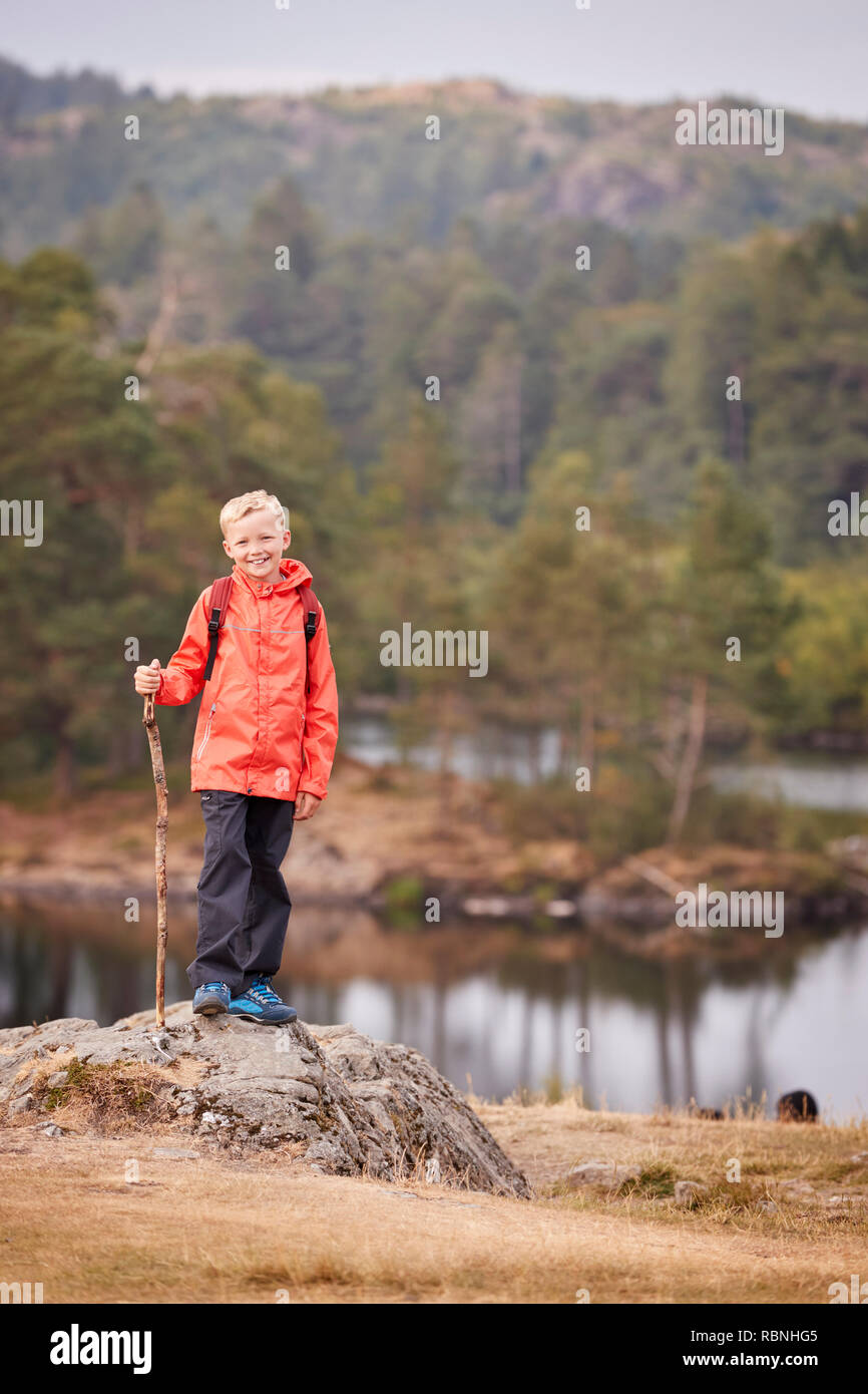 Ein Junge steht auf einem Felsen an einem See mit einem Stick, lächelnd auf Kamera, Lake District, UK, vertikal Stockfoto