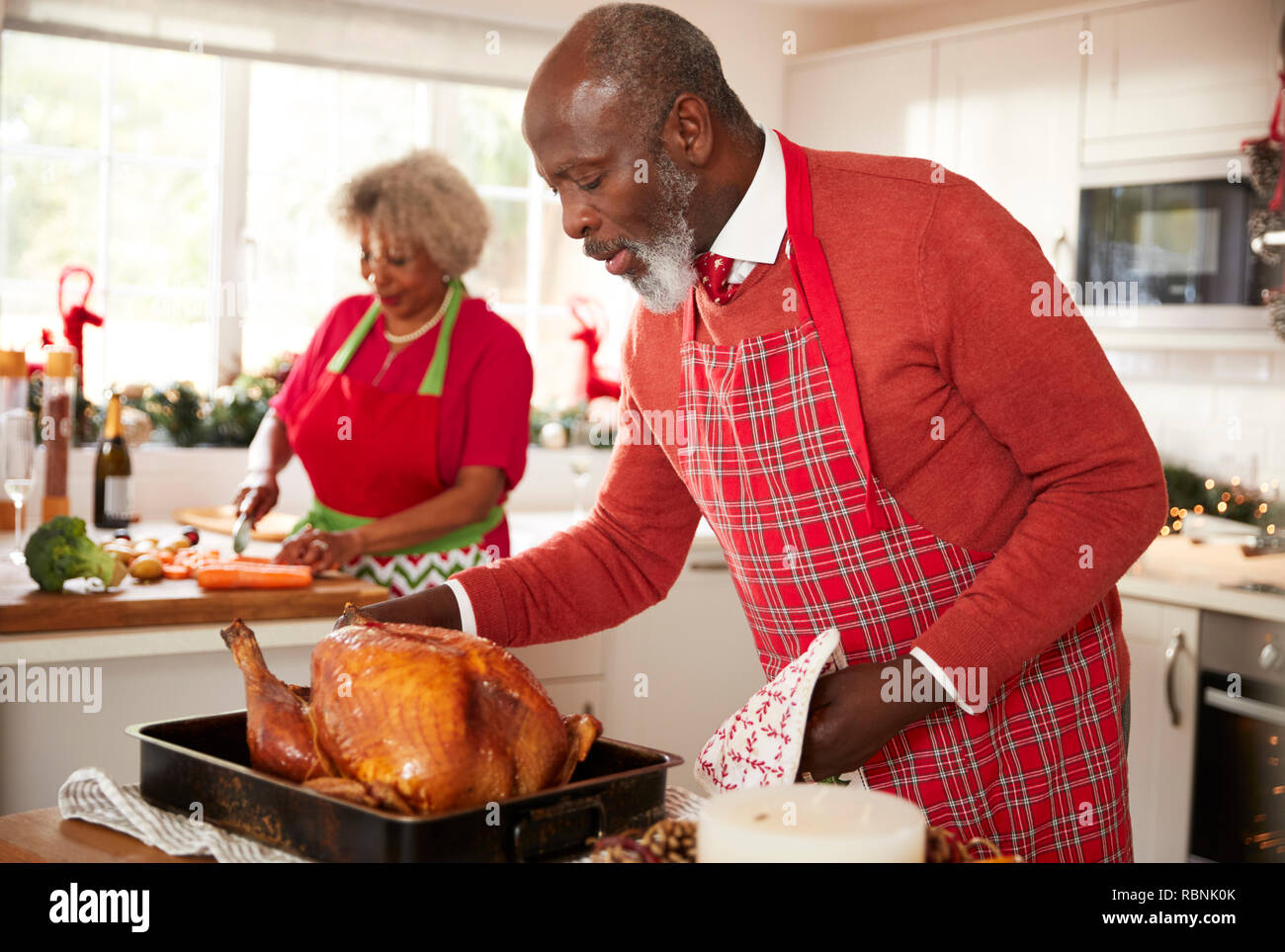 Älteren schwarzen Mann basting Truthahn in Vorbereitung für das Weihnachtsessen, seine Frau Zerkleinern von Gemüse im Hintergrund, in der Nähe Stockfoto