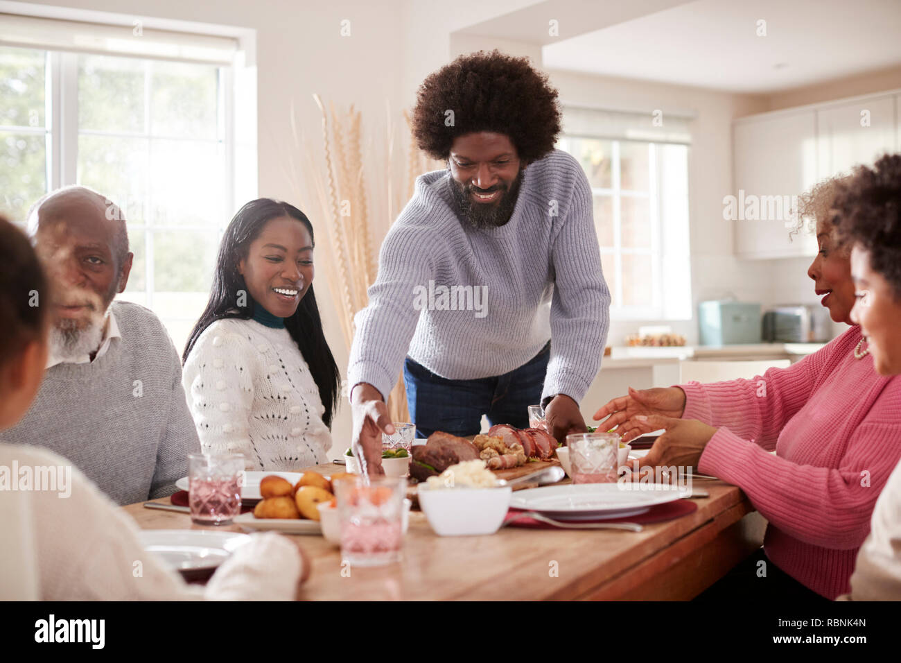 Im mittleren Alter schwarzer Mann bringen Braten auf den Tisch für den Sonntag Abendessen mit der Familie mit seinem Partner, Kinder und ihre Großeltern, Vorderansicht Stockfoto